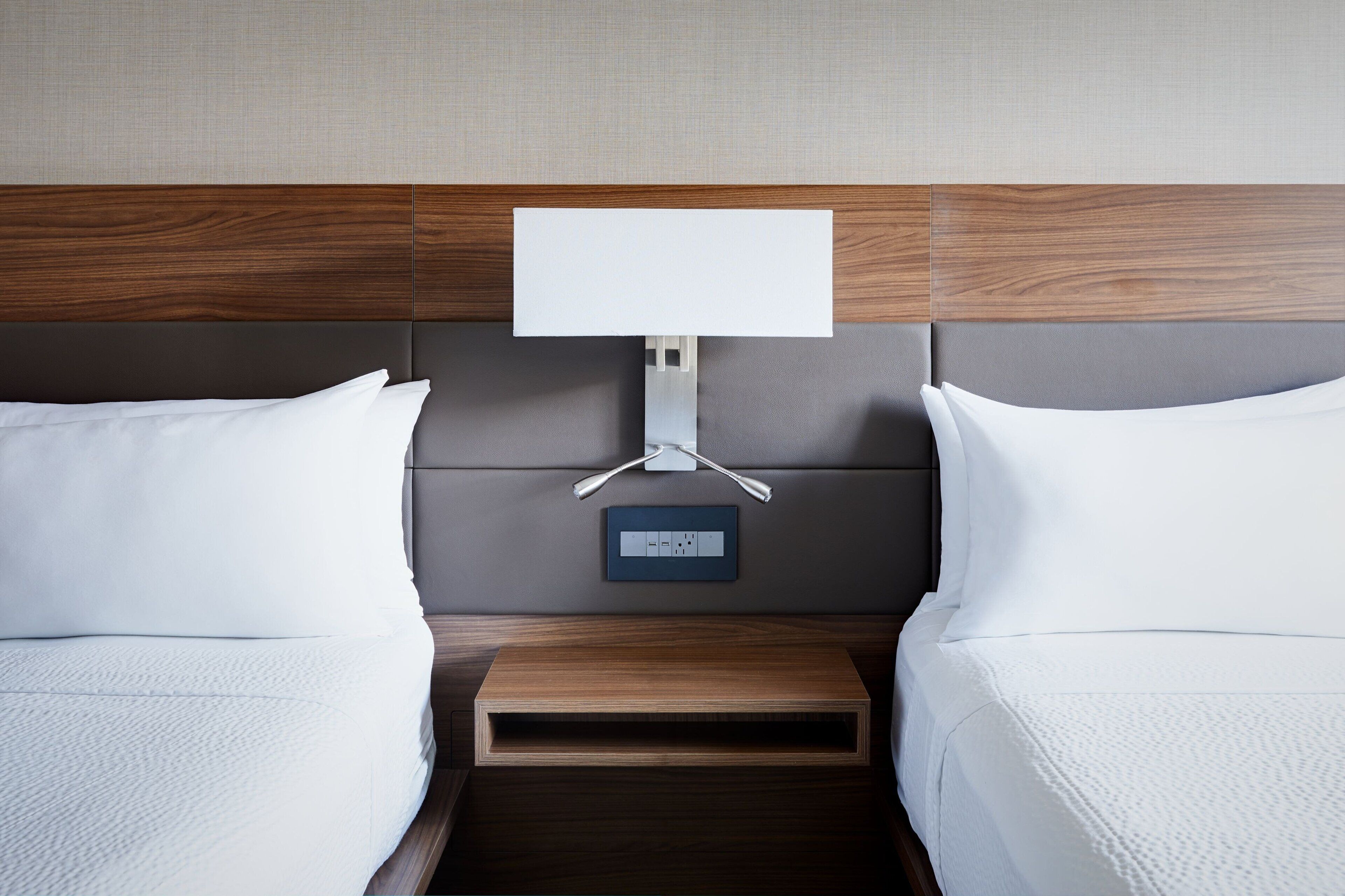 A hotel room with a wooden headboard, a bedside table with a power outlet, and two beds with white linens.