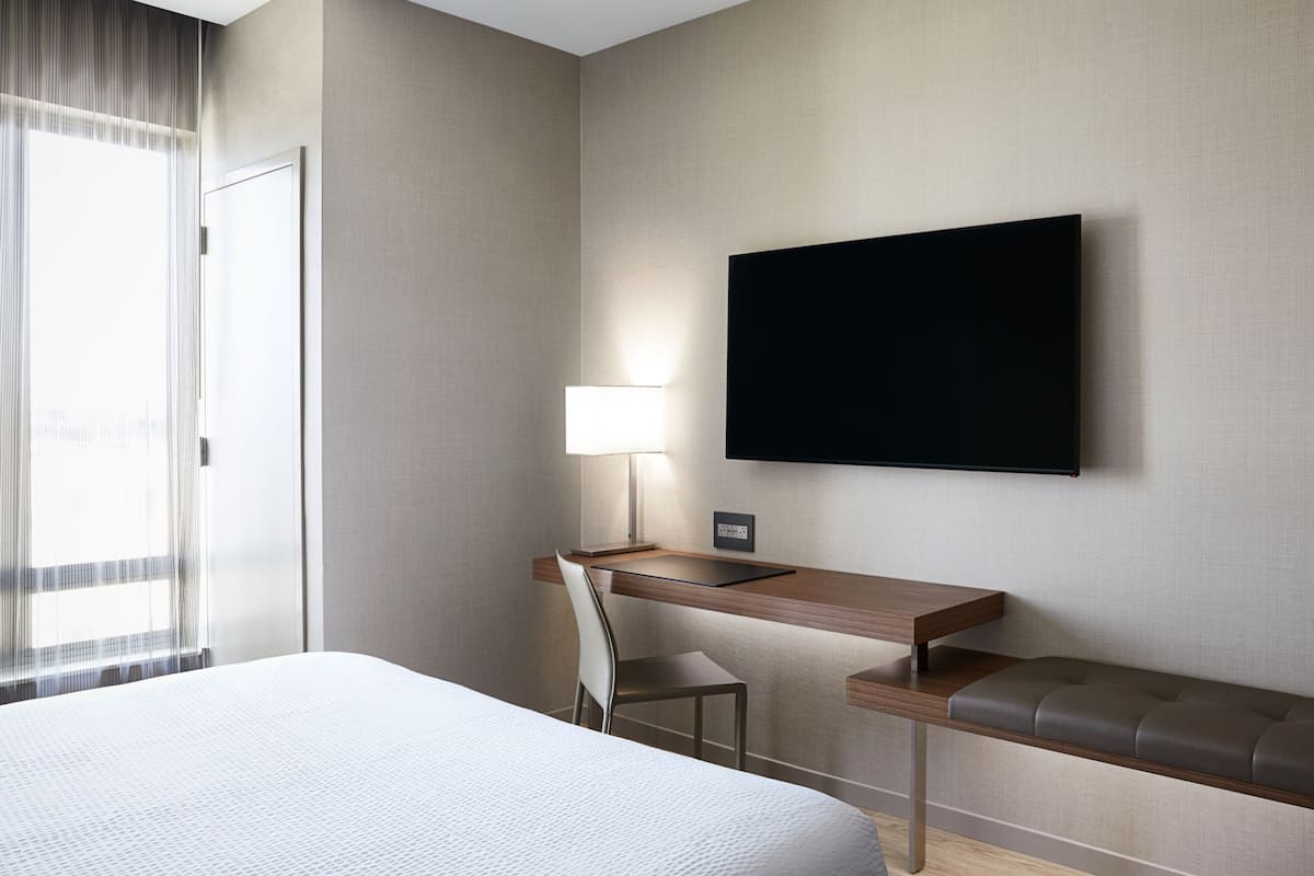 Habitación de hotel con una cama, un escritorio con silla, un televisor en la pared y una ventana con cortinas.