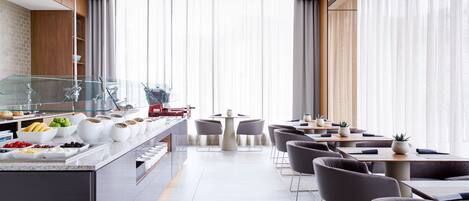 A modern restaurant with a long counter, various food items, and a seating area with tables and chairs.
