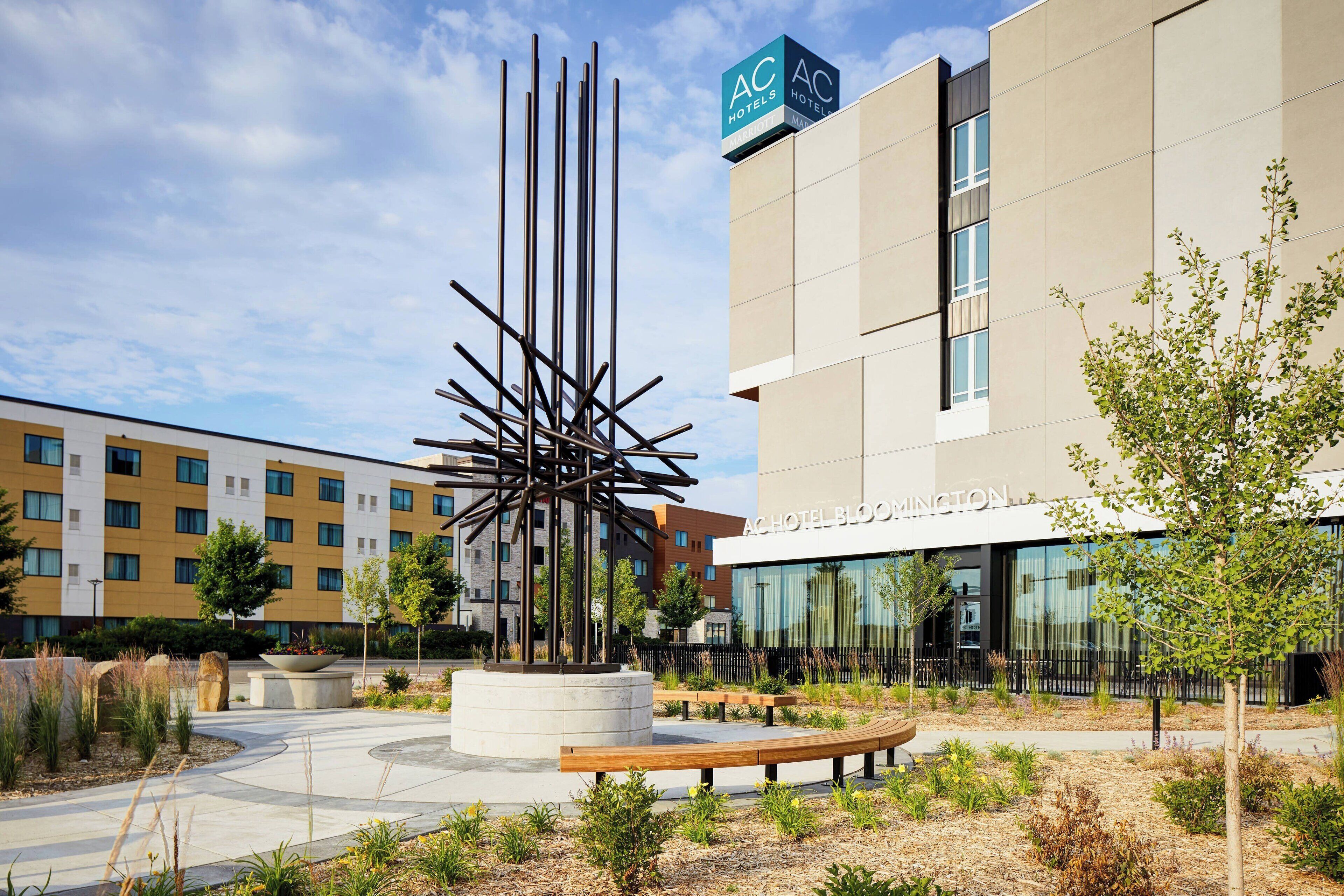 A modern hotel with a central sculpture, outdoor seating, and a well-maintained garden area.