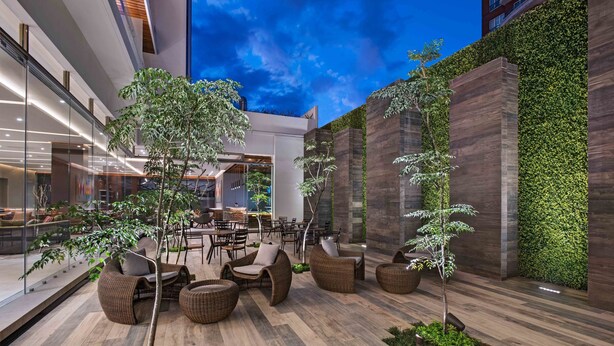 Un espacio moderno para sentarse al aire libre con mobiliario de mimbre y pequeños árboles, rodeado de paredes de vidrio y un suelo de madera.
