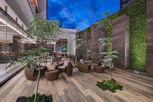 Un espacio moderno para sentarse al aire libre con mobiliario de mimbre y pequeños árboles, rodeado de paredes de vidrio y un suelo de madera.