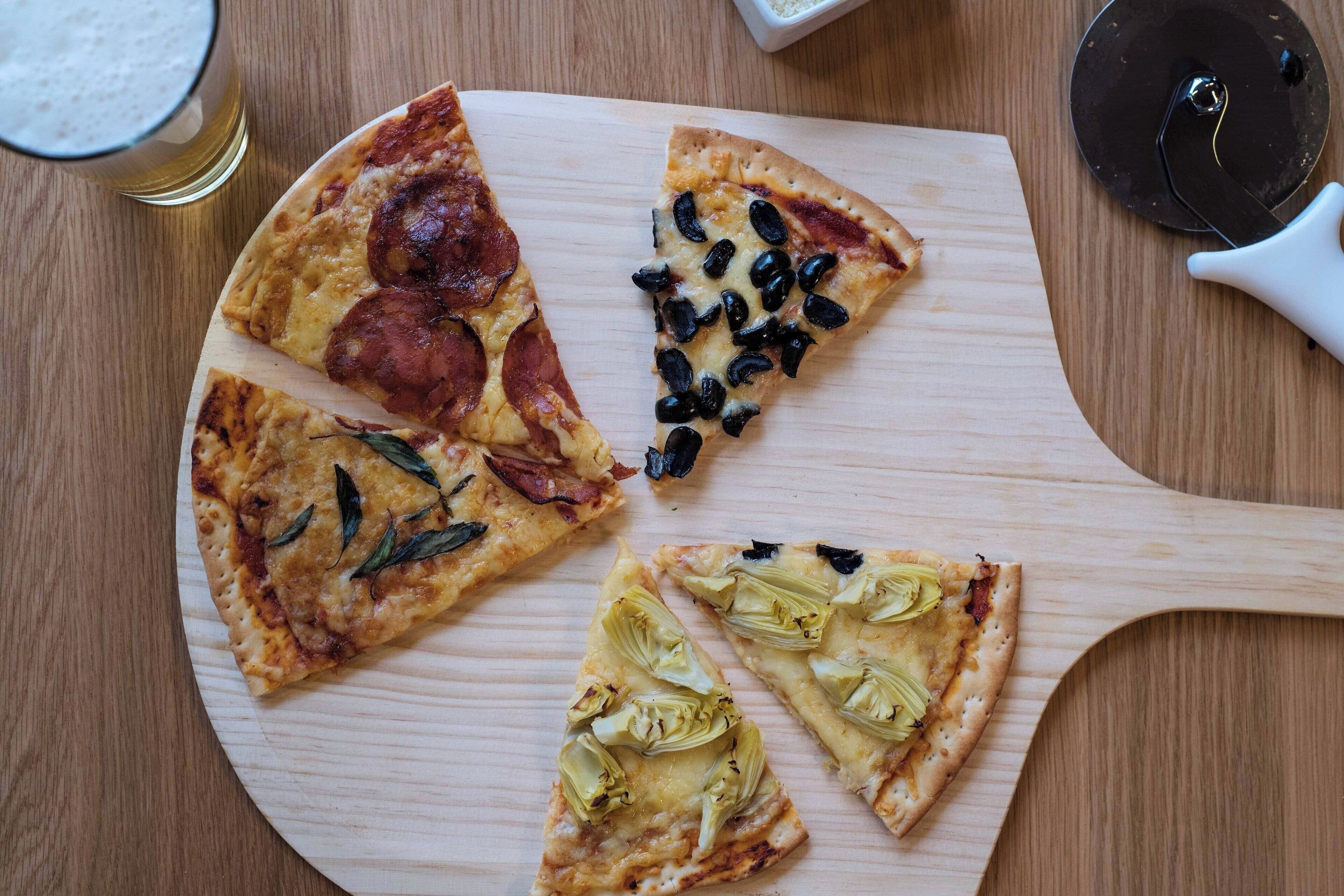 Un tagliere di legno con quattro fette di pizza, un tagliapizza e un bicchiere di birra su un tavolo di legno.