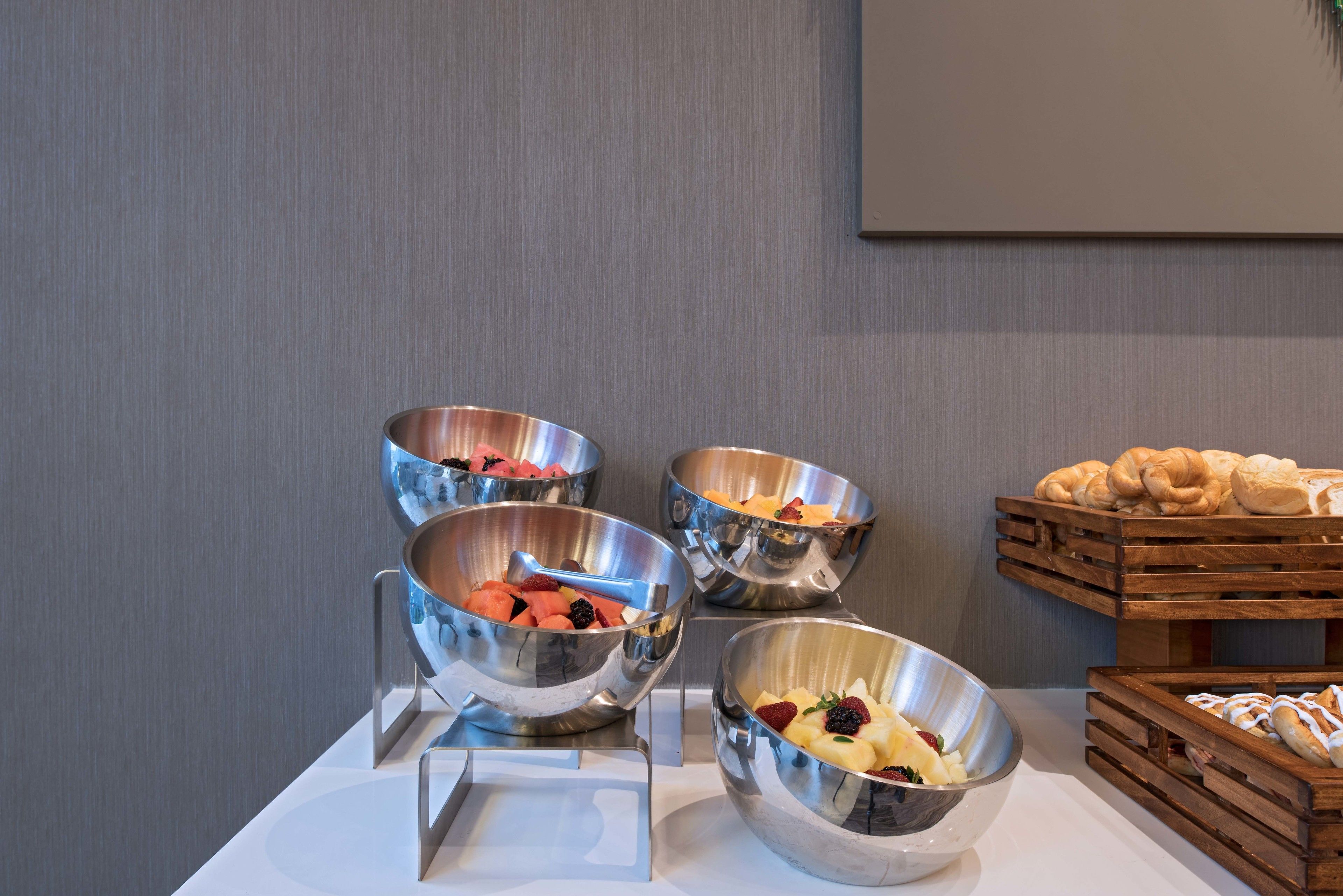 Ein Buffet mit vier Edelstahl-Schüsseln, gefüllt mit verschiedenen Früchten, und einer Holzkiste mit Gebäck.