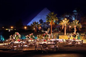 An outdoor dining area with round tables and chairs, decorated with flowers and illuminated by ambient lighting.