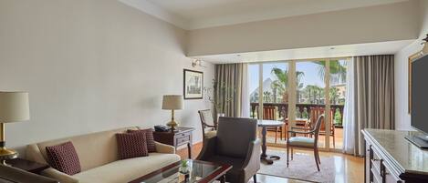 A spacious living room with a sofa, armchair, and coffee table, a television, and a balcony with a view of palm trees.
