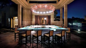 A well-lit outdoor bar area with a large rectangular table surrounded by chairs, a decorative ceiling light, and a view of a body of water in the background. - Marriott Mena House Cairo (Giza)