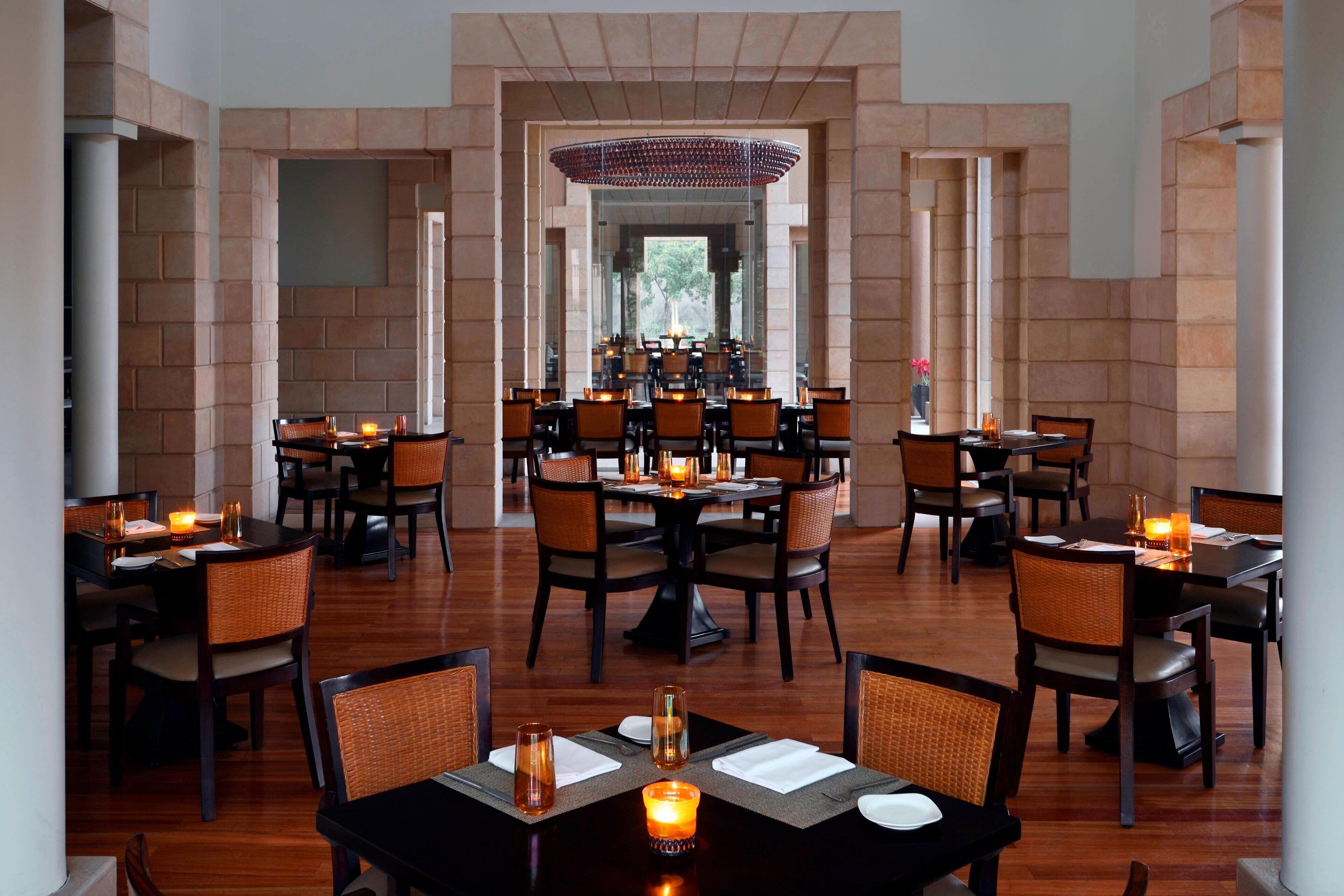 A restaurant with multiple tables and chairs, a central chandelier, and a brick wall backdrop.