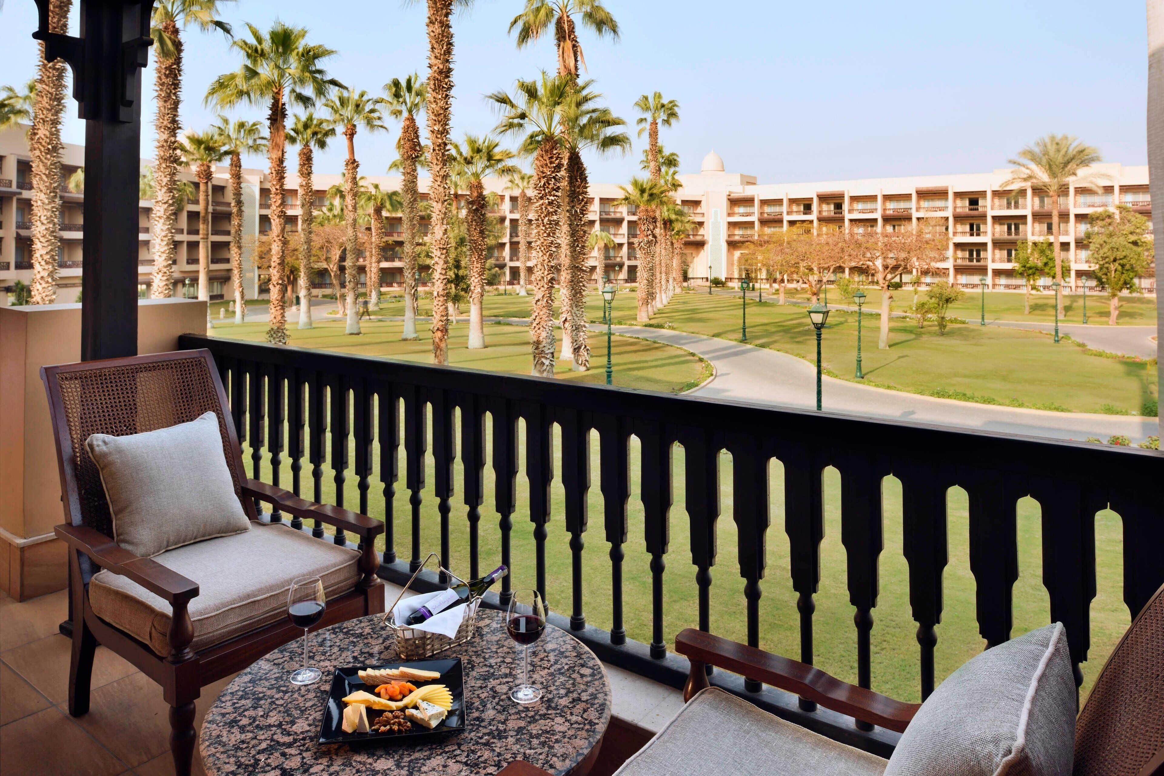 A balcony with a table set for two, featuring a bottle of wine, a glass, and a plate of snacks. The balcony overlooks a well-maintained lawn with palm trees and a building in the background.