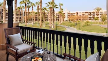 A balcony with a table set for two, featuring a bottle of wine, a glass, and a plate of snacks. The balcony overlooks a well-maintained lawn with palm trees and a building in the background.
