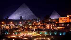 A nighttime view of a resort with two illuminated pyramids in the background, a pool area with lights, and palm trees. - Marriott Mena House Cairo (Giza)