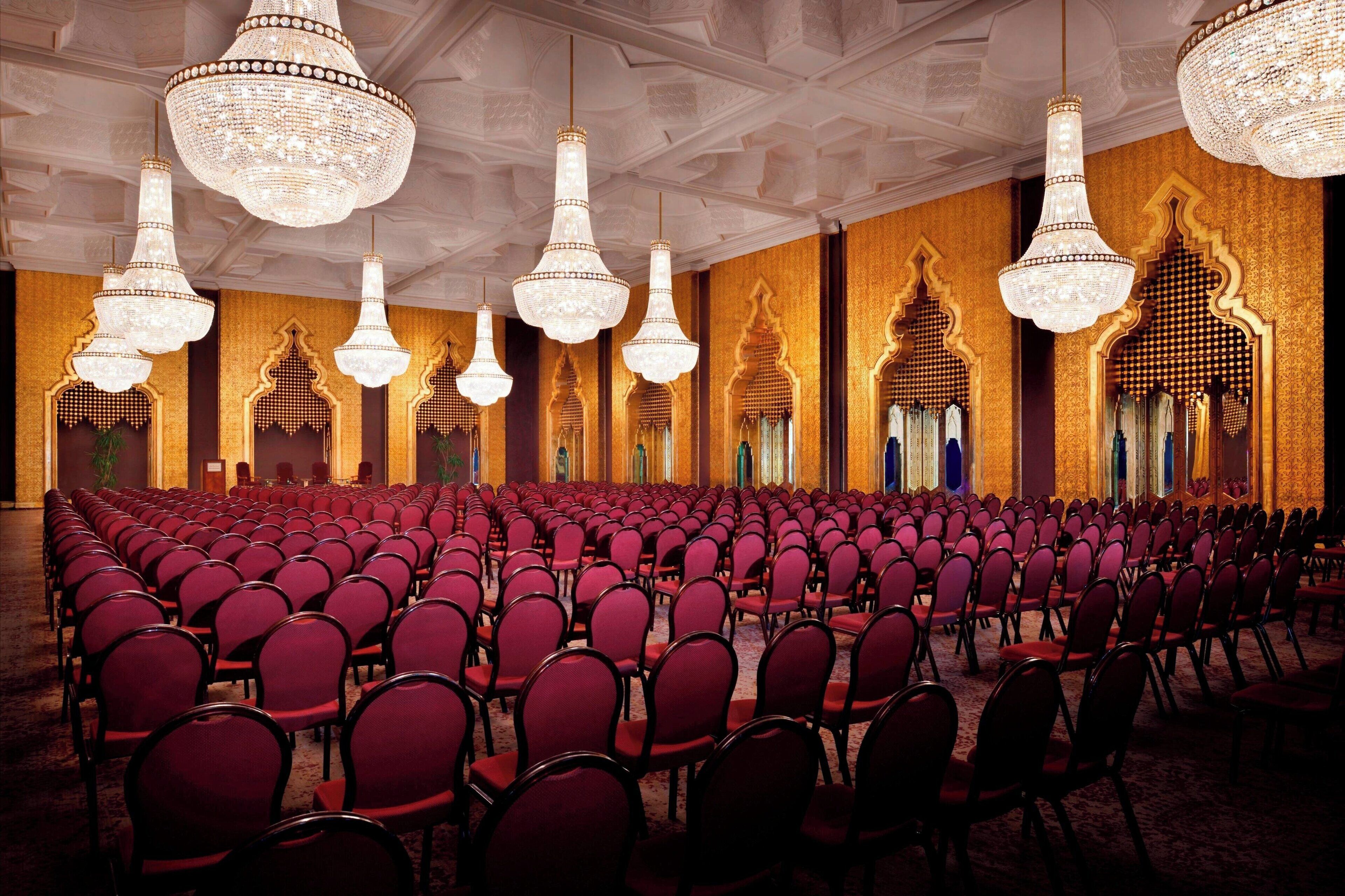 A conference room with rows of red chairs, ornate chandeliers, and detailed wall decorations.