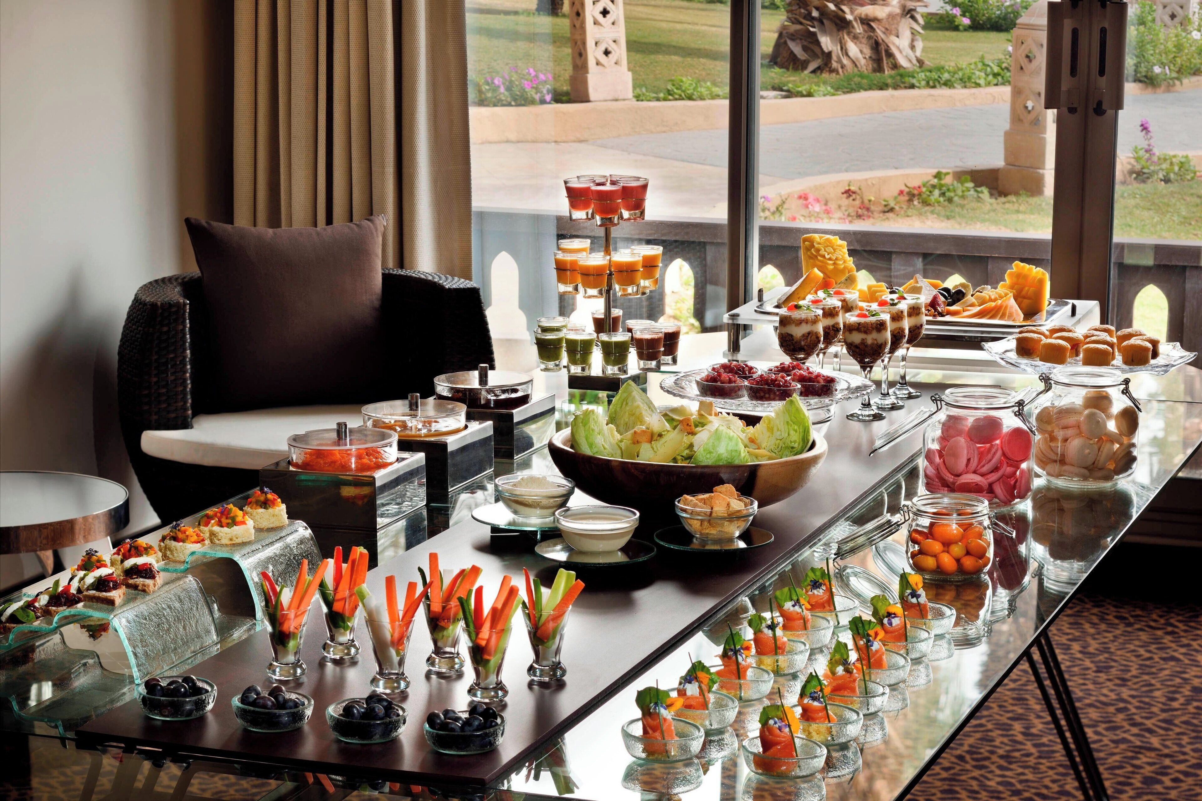 Una mesa de buffet con diversos alimentos y bebidas, un sofá, y vistas al exterior.