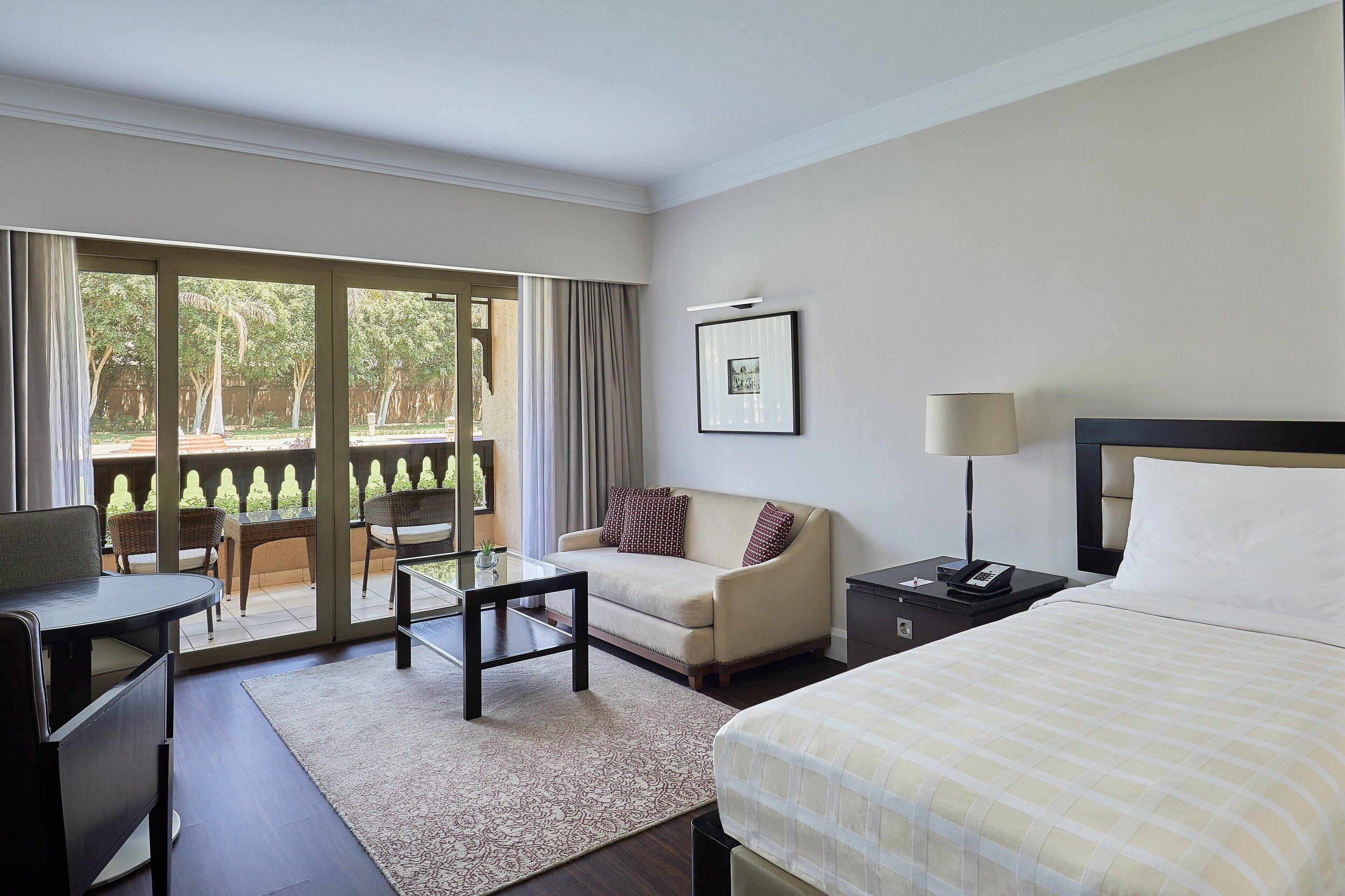 A hotel room with a bed, a sofa, a desk, and a balcony with outdoor furniture.