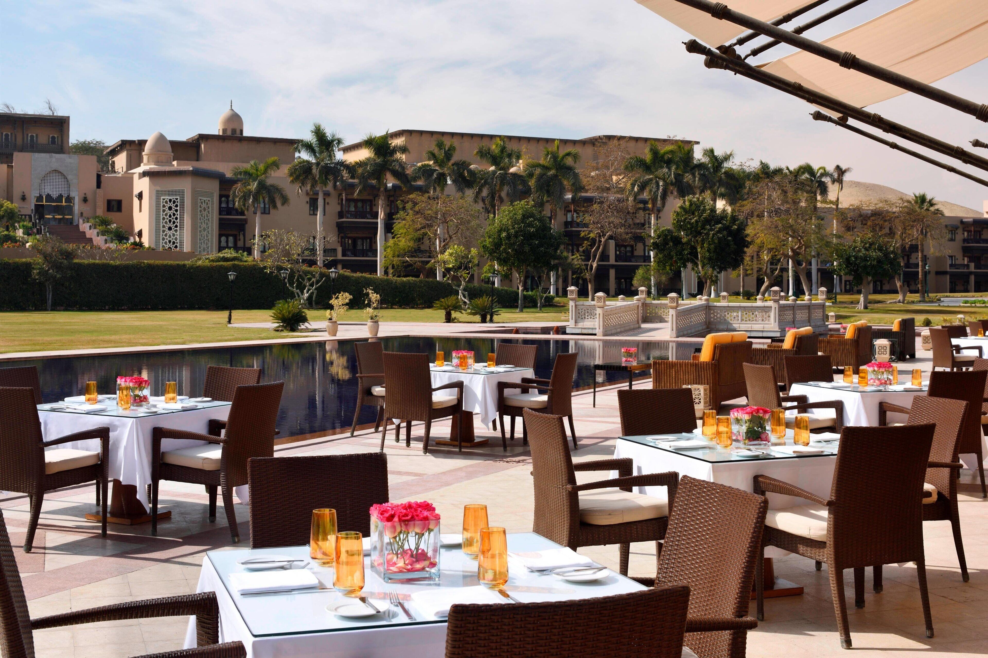 An outdoor dining area with tables set for two, surrounded by palm trees and a pool.