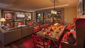 A dining area with a long wooden table set for a meal, surrounded by red chairs. A fireplace is visible in the background, and there is a sofa and a television in the adjacent living area. - Heavenly Peak 310 4 BedroomCondo By Moving Mountains (Steamboat Springs)