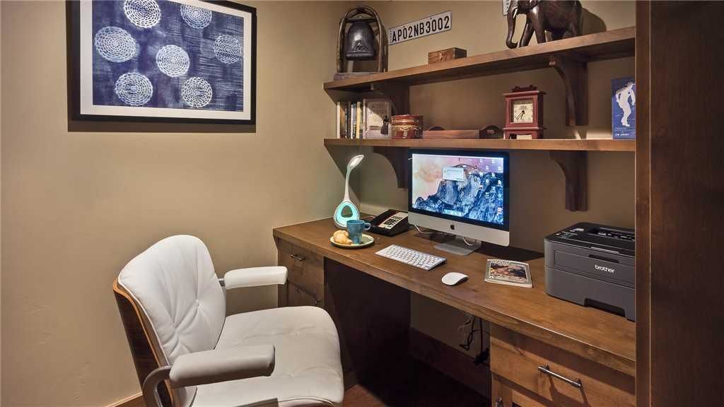 A home office with a desk, computer, printer, and a chair. There is a framed artwork on the wall and a shelf with various items.