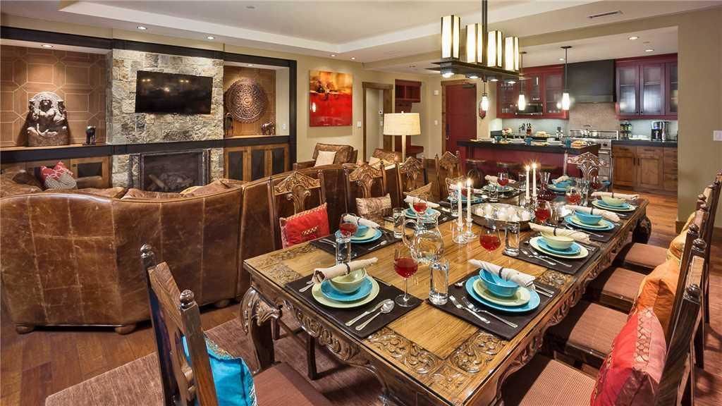 A dining area with a long table set for six, surrounded by upholstered chairs. A fireplace is visible in the background, and a television is mounted on the wall.