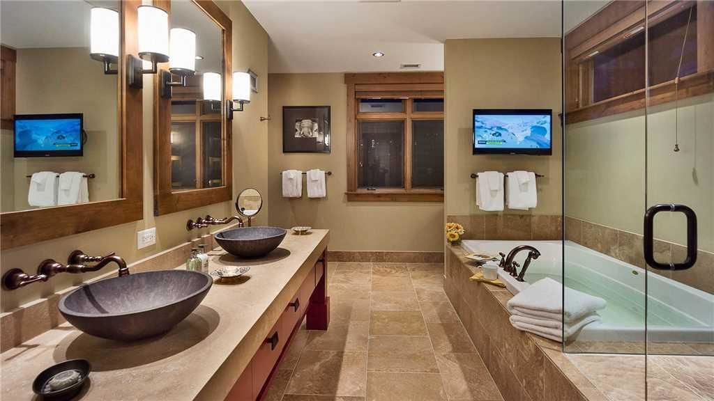 A modern bathroom with a large mirror, two sinks, a bathtub, and a television.