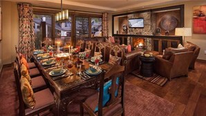 A dining area with a long wooden table set for a meal, surrounded by leather chairs. A fireplace is visible in the background, and a television is mounted on the wall. - Zen Mountain 311 3 BedroomCondo By Moving Mountains (Steamboat Springs)