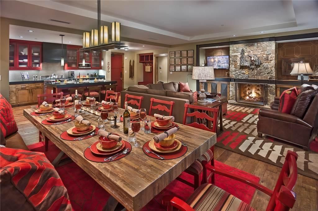 Un coin repas avec une longue table en bois dressée pour le repas, des chaises rouges et une cheminée en arrière-plan.