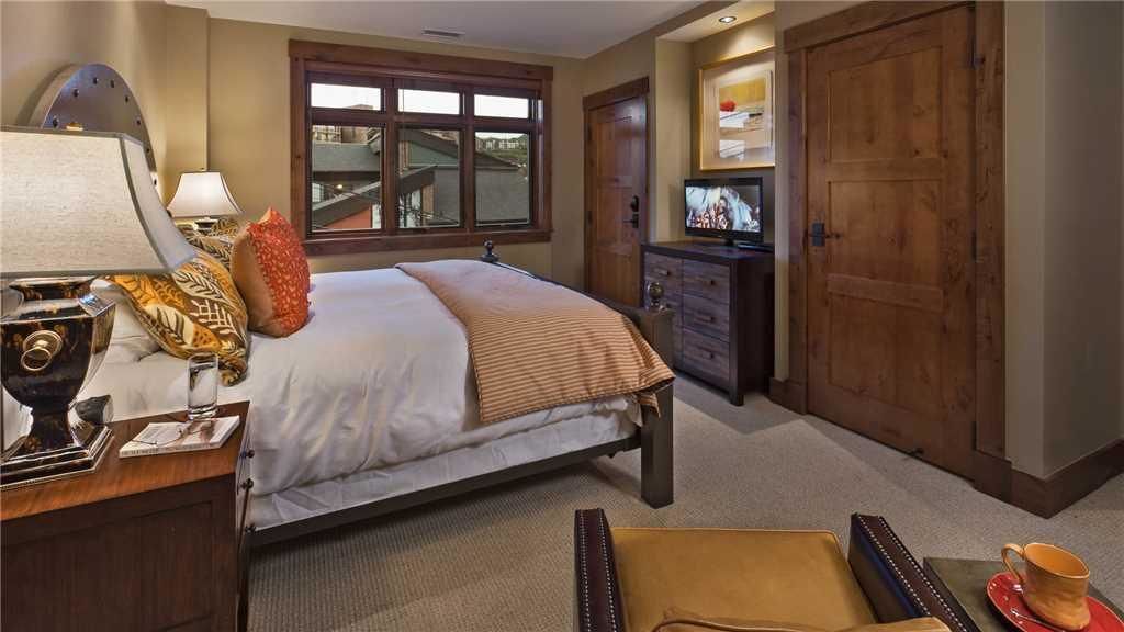 A hotel room with a bed, a desk with a lamp, a television, a wooden wardrobe, and a large window.