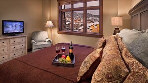 A hotel room with a bed, a TV, a window with a view of a town, a tray with fruits and wine, and a lamp. - Sawtooth Mountain 514 4 BedroomCondo By Moving Mountains (Steamboat Springs)