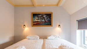 Uma cama de casal com roupa de cama branca e duas almofadas, um quadro de uma árvore na parede e duas luzes fixadas na parede.