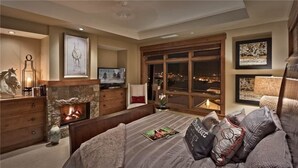 Quarto com lareira de pedra, televisão, janela ampla com vista noturna e cama com muitas almofadas.