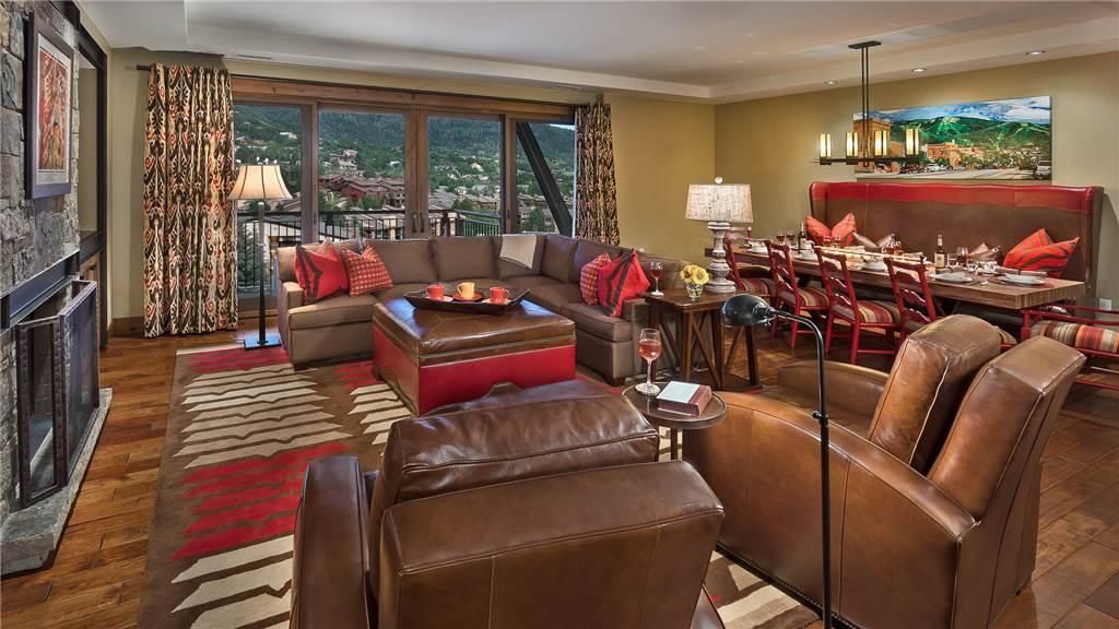 A spacious living room with a fireplace, a sofa set, a dining area with red chairs, and a view of the mountains through large windows.