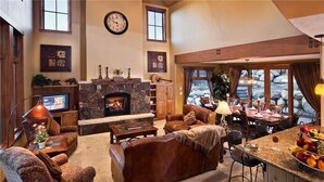 A spacious living room with a fireplace, leather sofas, a dining area with a stone fireplace, and a large window overlooking a rocky landscape.
