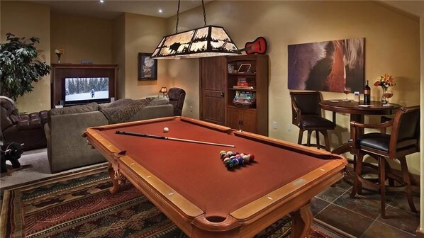 Una mesa de billar en una sala de estar con un televisor, una lámpara y un comedor con sillas.