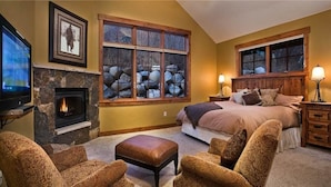 A bedroom with a fireplace, large windows, a bed, and a TV.