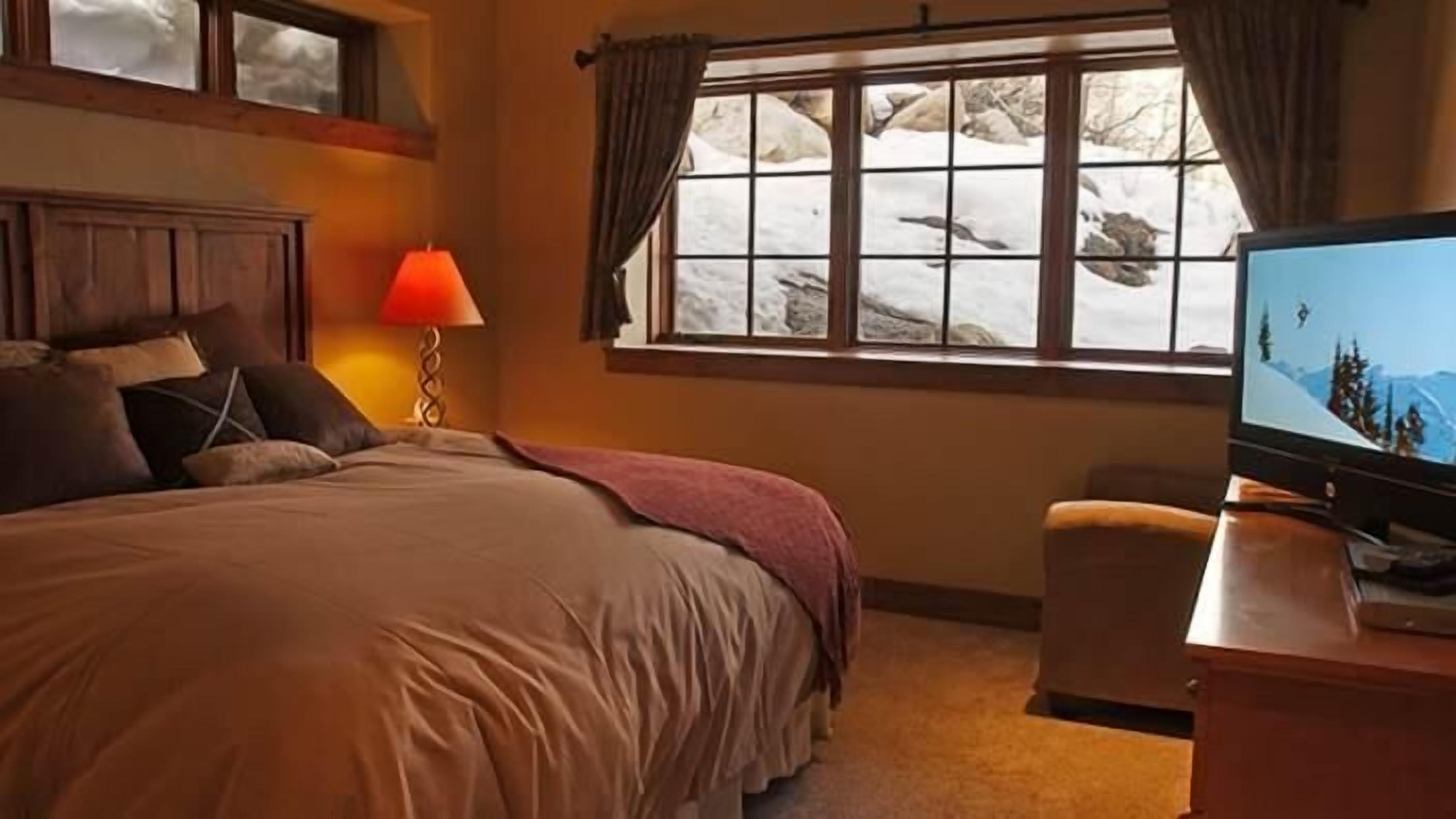Een slaapkamer met een groot bed, een televisie op een houten standaard, een raam met uitzicht op besneeuwde bergen en een lampje op een nachtkastje.