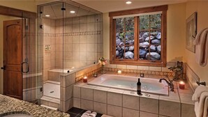 A bathroom with a large bathtub, a glass-enclosed shower, a window with a view of rocks, and a wooden door.