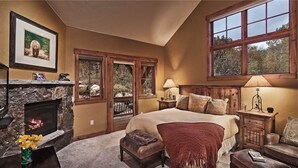 A bedroom with a stone fireplace, a large bed with multiple pillows, a nightstand, and a window with a view of trees.