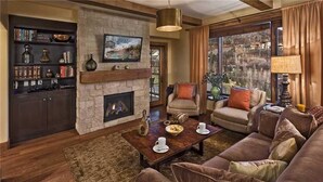 A cozy living room with a fireplace, comfortable seating, a bookshelf, and a large window offering a scenic view.