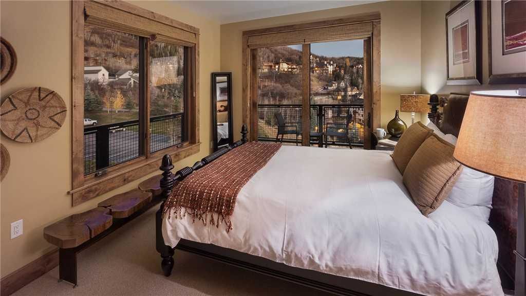 A bedroom with a large bed, wooden bench, and a view of a town through large windows.