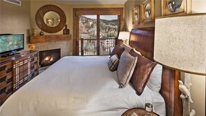 A large bed with multiple pillows, a television on a wooden dresser, a fireplace, and a view of a town through a balcony door.