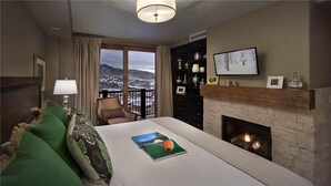 A hotel room with a bed, a fireplace, a television, and a view of a snowy landscape through the window. - Skyfall Ridge 702 2 BedroomCondo By Moving Mountains (Steamboat Springs)