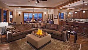 A spacious living room with a large sectional sofa, a coffee table with candles, and a view of a cityscape through large windows.