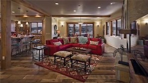 A spacious living room with a large red sectional sofa, a coffee table, and a patterned rug. There are multiple windows offering a view of the mountains.