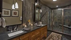 A modern bathroom with a large mirror, two sinks, wooden cabinets, and a shower area with stone walls.