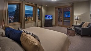 A bedroom with a large bed, a television, a chair, and a view of a cityscape through the windows.