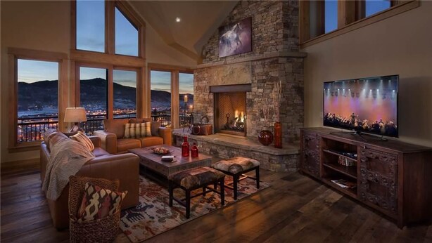A spacious living room with a stone fireplace, a large flat-screen TV, and a view of a cityscape through large windows.