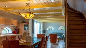 Uma sala de jantar ampla, com mesa e cadeiras de madeira, cozinha ao fundo e escada que leva a um andar superior.