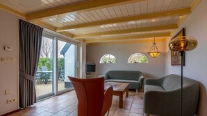 Sala de estar com sofá, poltrona e uma mesa. Possui uma porta de vidro deslizante que dá para uma área externa.