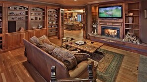 Un salon spacieux avec un grand canapé, une cheminée, un téléviseur et des étagères en bois où sont exposés divers objets.