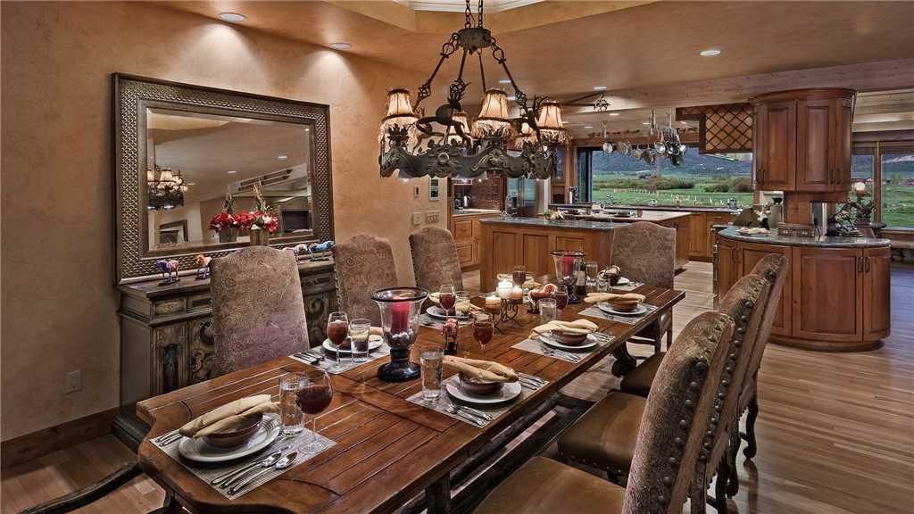 A dining room with a long wooden table set for a meal, surrounded by upholstered chairs. A large mirror and chandelier are visible on the walls.