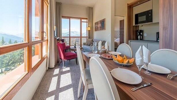 Comedor con una mesa dispuesta para dos, cocina al fondo y vista a las montañas a través de amplios ventanales.